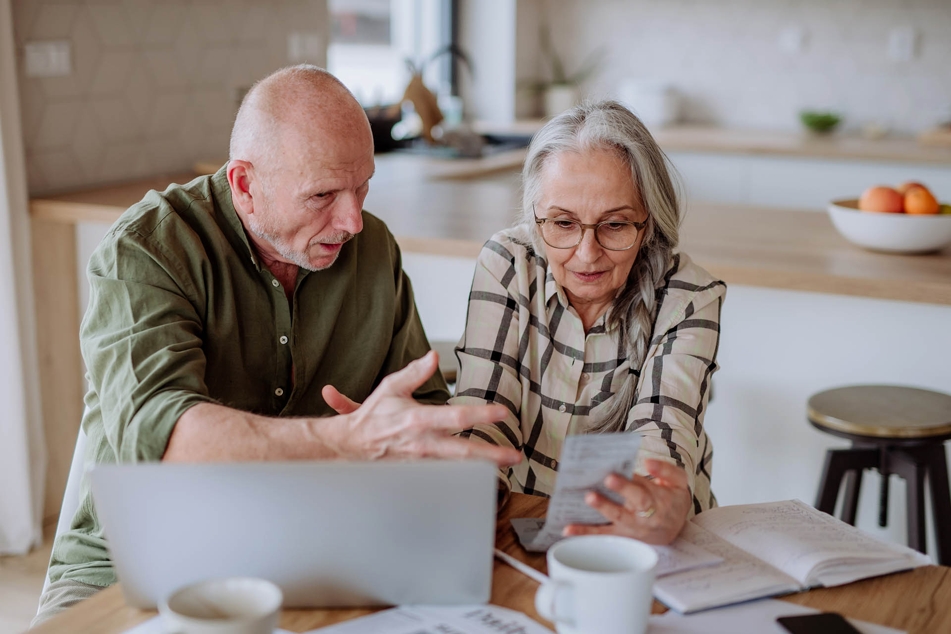 mature couple intently discussing receipts at kitchen table near computer